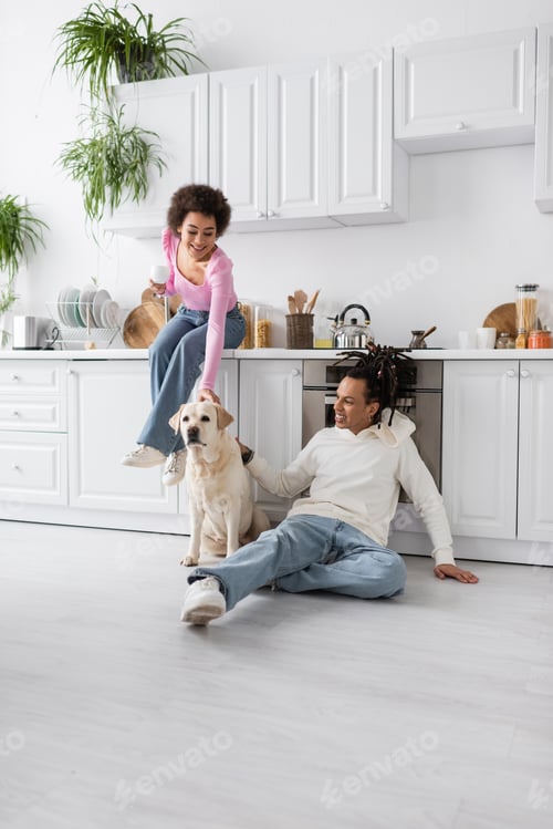 Preview: Cheerful african american woman holding coffee near boyfriend and labrador on floor in kitchen