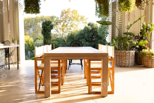Preview: Dining table with wooden chairs in sunlit patio, surrounded by lush greenery
