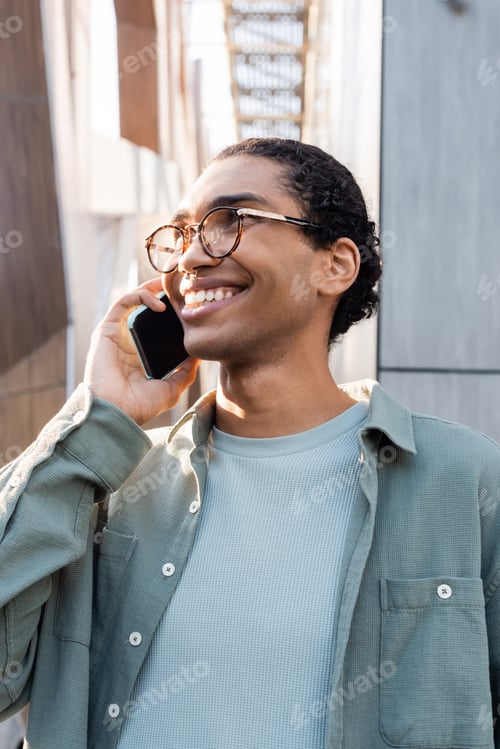 Preview: joyful african american man in grey shirt and eyeglasses talking on smartphone on urban street