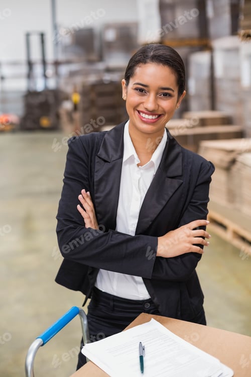 Preview: Woman wearing blazer standing with crossed arms in warehouse beside wheeled cart holding paperwork