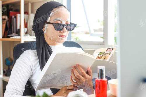 Preview: Young Asian Muslim Woman wearing sunglasses with facial sheet mask applied relaxing at home and read