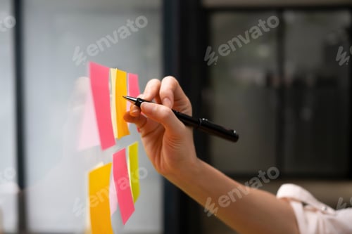 Preview: Close up view businesswoman writing creative ideas on sticky notes on glass wall.