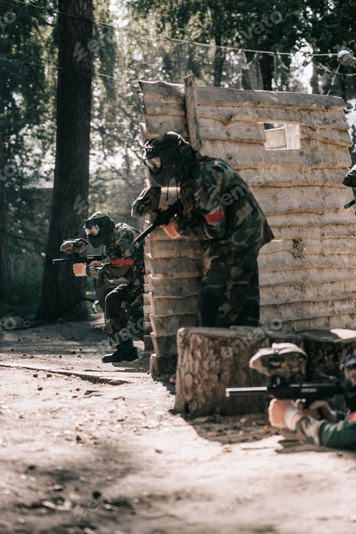Preview: paintball team in uniform and protective masks playing paintball with marker guns outdoors