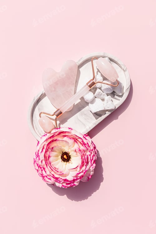 Preview: Rose quartz roller massager and gua sha scraper on a white ceramic tray with a delicate pink flower.