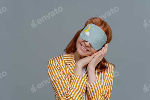 Preview: Smiling gentle woman with eye mask, tilts head on pressed palms, wears striped pajama