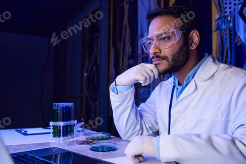 Preview: indian scientist in goggles working on laptop near test tubes and petri dishes in futuristic lab