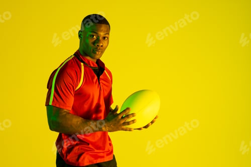 Preview: Portrait of african american male rugby player with rugby ball over yellow lighting