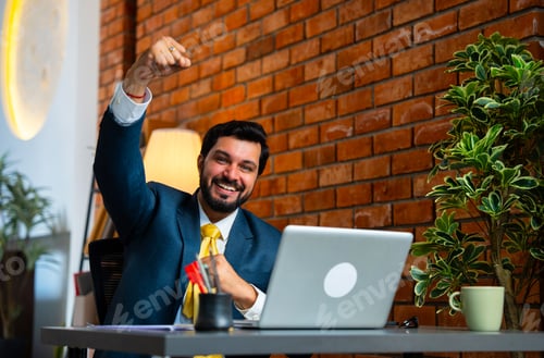 Preview: Smiling Indian businessman celebrates a big win while working on laptop in cozy office setup