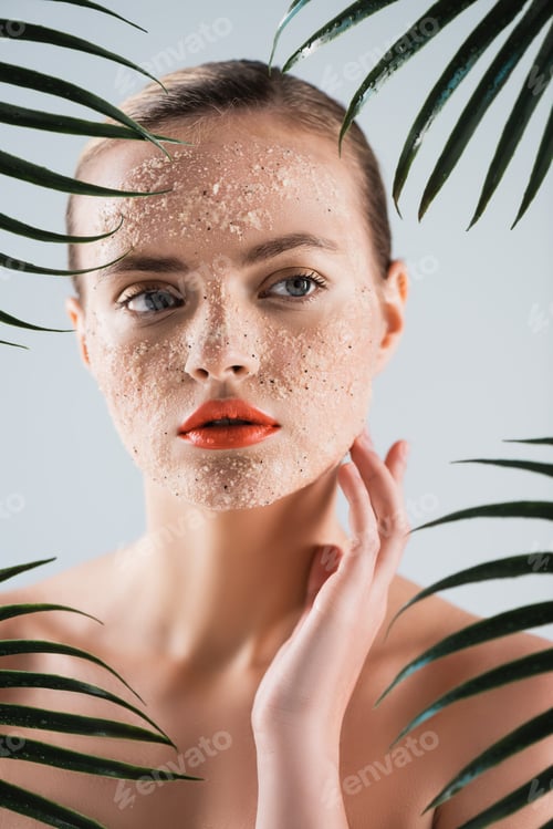 Preview: selective focus of naked woman with scrub touching face near palm leaves on white