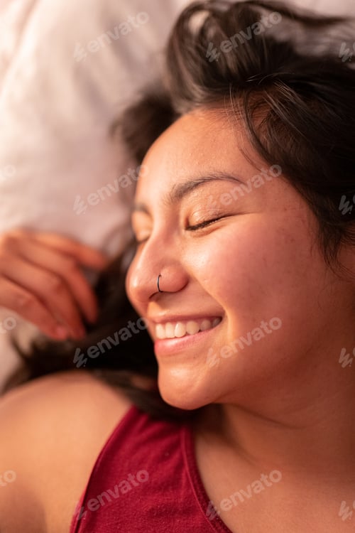 Preview: A young woman wearing a piercing is lying on bed happily smiling before sleep. Close up