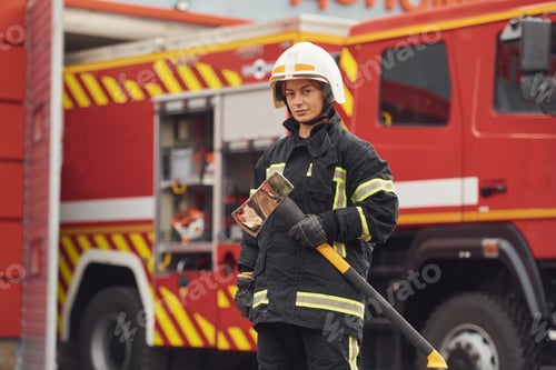 Preview: Standing with axe in hands against truck. Woman firefighter in uniform is at work in department