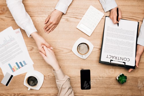 Preview: Top view of business partners shaking hands near contract, coffee and credit card on table