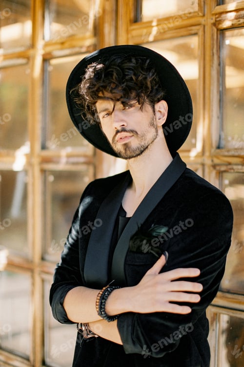Preview: Young man in a black hat stands with his arms crossed against the background of a wooden window