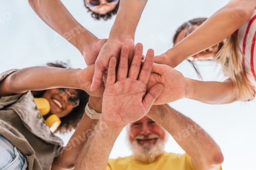 Preview: Low angle of cheerful friends in casual clothes stacking hands outside and looking down while