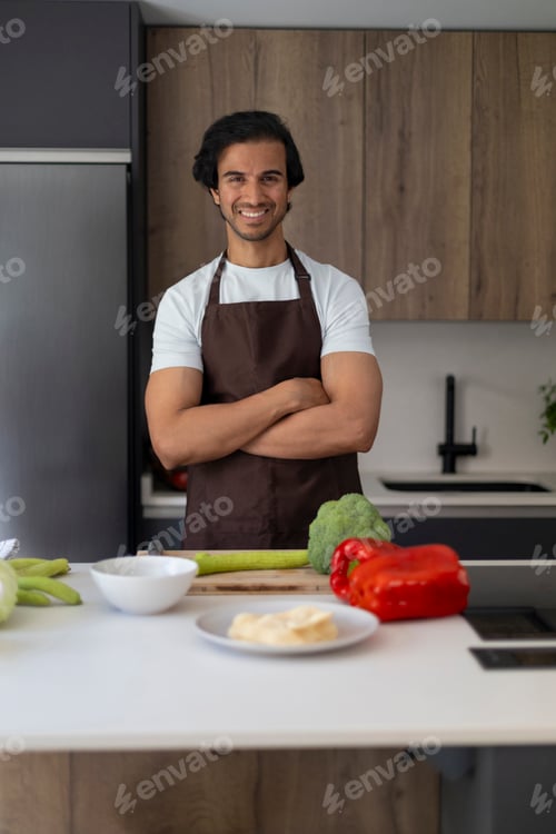 Preview: Smiling chef with crossed arms in a modern kitchen surrounded by fresh vegetables.