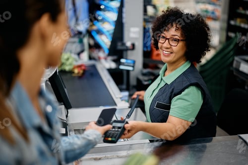 Preview: Happy cashier holding credit card reader while customer is paying with smart phone at supermarket.