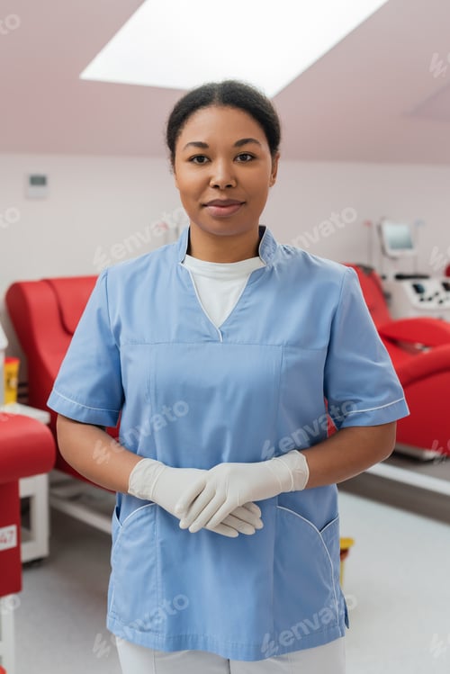 Preview: positive multiracial nurse in blue uniform and latex gloves looking at camera near medical chairs
