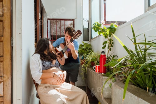 Preview: Latin young couple of man singing and playing guitar with his pregnant wife, at home