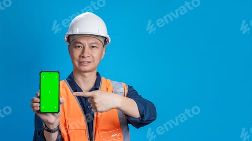 Preview: Confident young engineer wearing orange and a safety helmet stands holding a laptop, phone