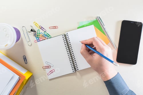 Preview: View of a male hand writing in a notebook, with coffee cup, folders, supplies and smartphone.
