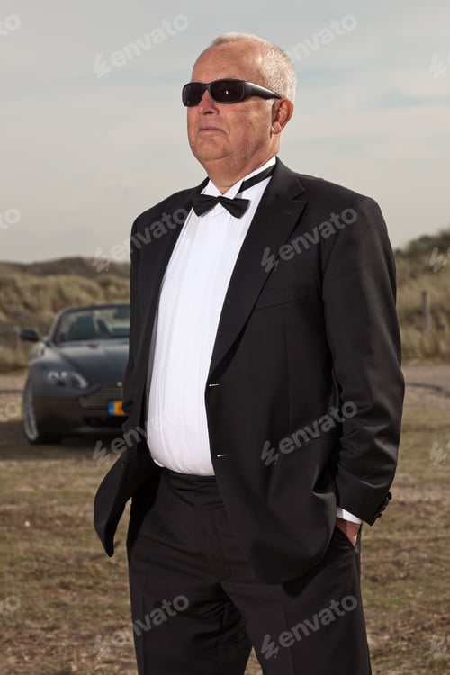 Preview: Senior Good Looking Happy Relaxed Retired Man With His Sports Car In Dune Landscape With Cloudy
