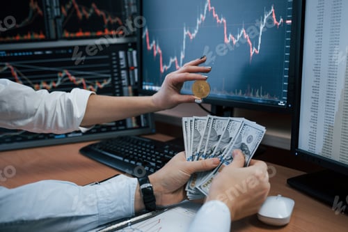 Preview: Woman's and man's hand holding bitcoin and money in modern office with many monitors with graphs
