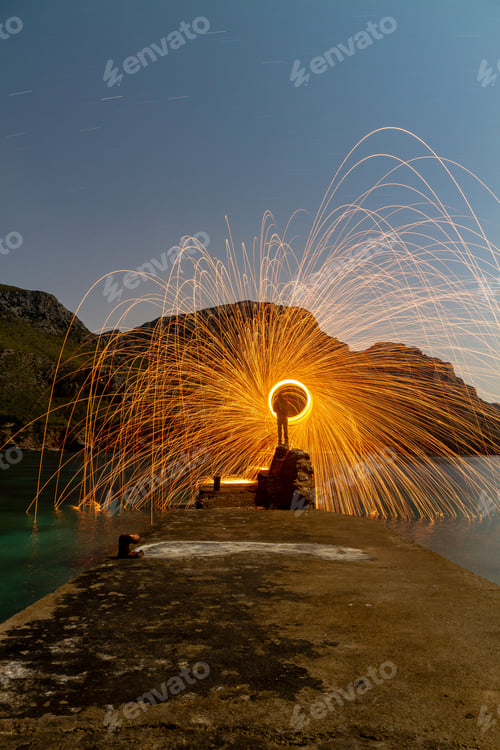 Preview: Person with burning circle on stones between water near hills at night