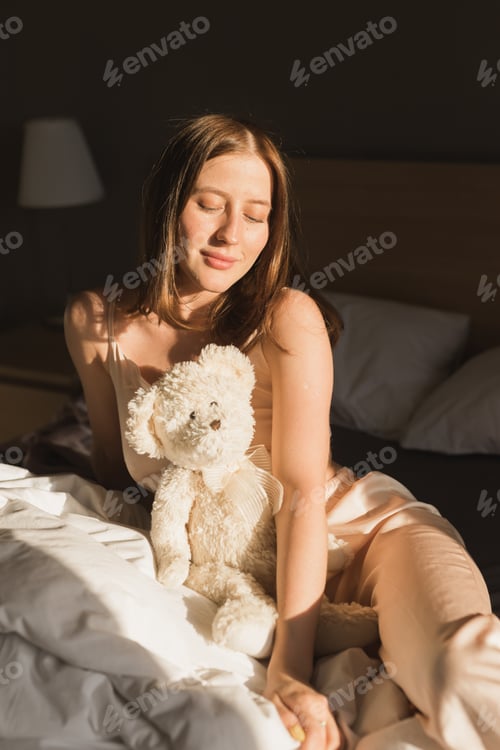Preview: Portrait woman waking up in her bed, she is smiling and stretching with toy bear. Happy young woman