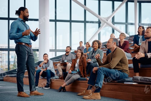 Preview: Black businessman talking to large group of entrepreneurs during education event in conference hall.
