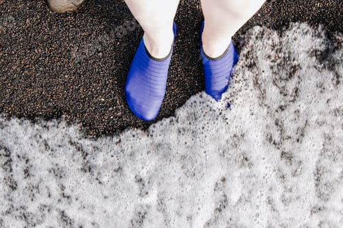 Preview: Woman's feet in underwater shoes on a black beach with foaming waves.