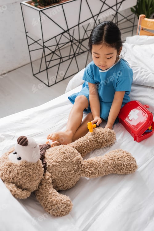 Preview: high angle view of asian child playing on hospital bed and examining teddy bear with toy reflex