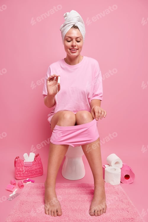 Preview: Happy woman sits on toilet holds tampon has menses cares about hygiene wears wrapped towel over head