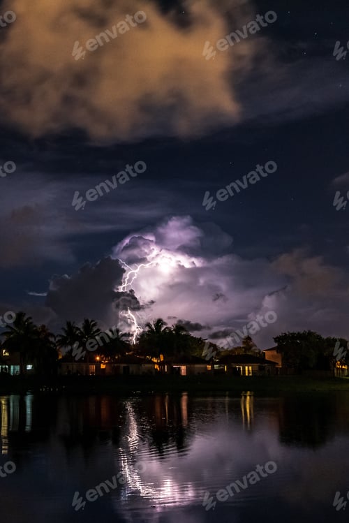 Preview: Lightning amongst the storm clouds is reflected in a lake behind houses with lights on.