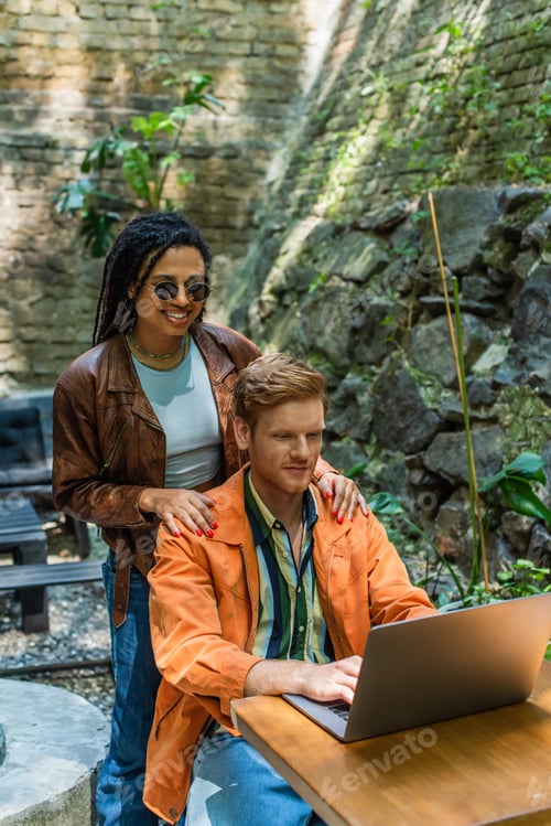Preview: happy african american woman touching shoulders of redhead man using laptop in outdoor terrace