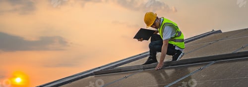 Preview: man in a yellow safety vest is looking at a laptop while standing on a roof