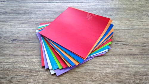 Preview: Top-down view of a stack of multicolored origami paper on a wooden background.