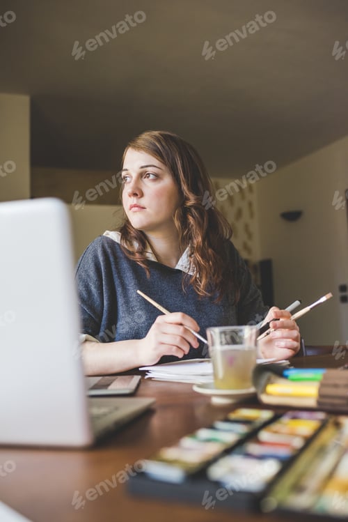 Preview: Young caucasian woman artist sitting at desk holding brushes