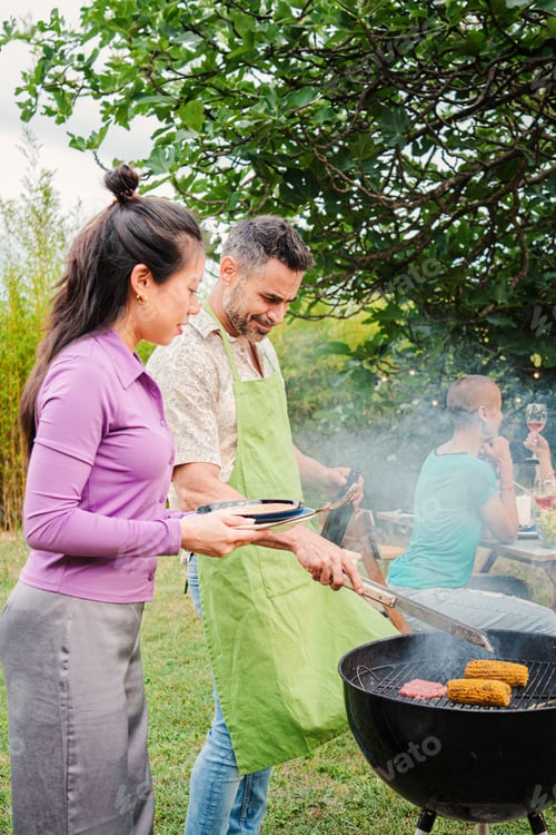 Preview: Vertical portrait. Middle adult happy couple cooking meat on a barbecue bbq party with their friends