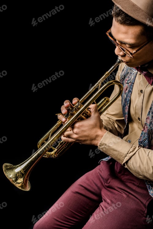 Preview: mixed race male jazzman in hat and eyeglasses playing on trumpet isolated on black