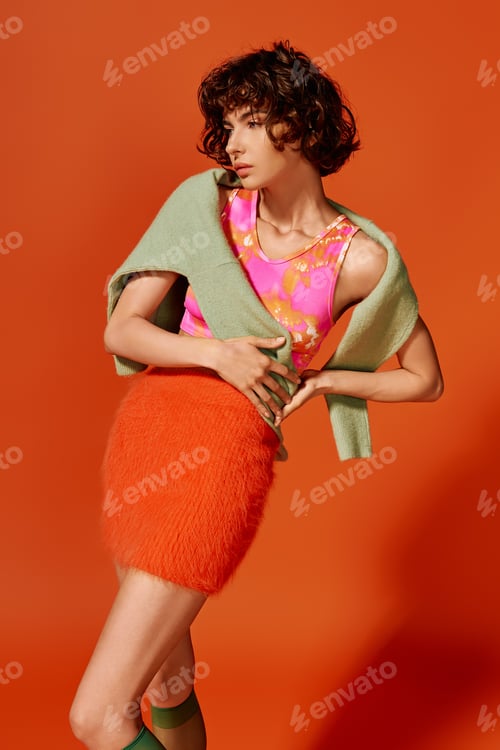 Preview: Emotional young woman poses gracefully in vibrant outfit against an orange backdrop