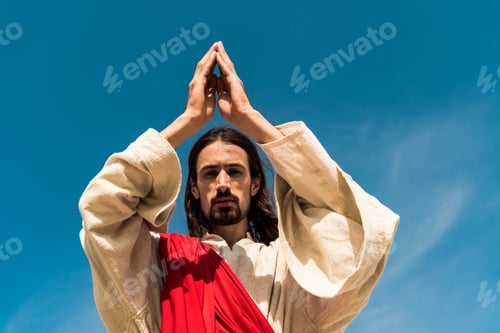 Preview: low angle view of jesus with praying and looking at camera against blue sky