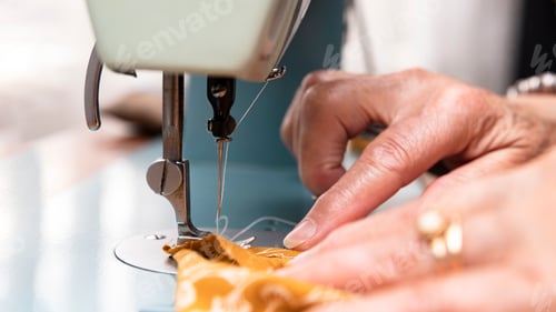 Preview: Hands of older woman sewing an orange cloth on a sewing machine in the foreground