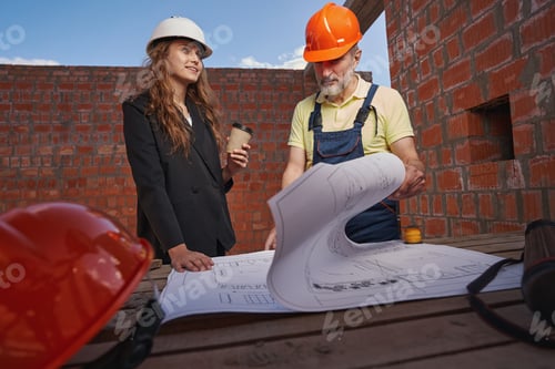 Preview: Woman engineer looking to side while worker studying blueprints