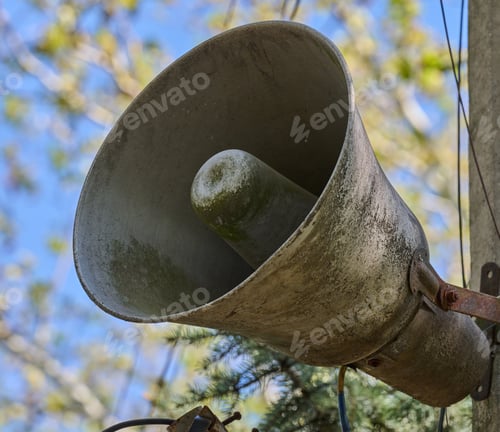 Preview: An old, dirty, grey loudspeaker is mounted on a pole. In the background