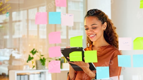 Preview: Happy female designer planning ideas on a glass wall with colorful sticky notes inside a creative a