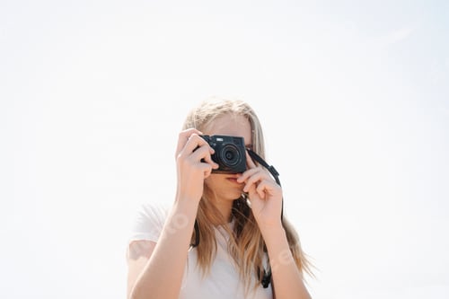 Preview: Teenage girl with long blond hair looking through a camera, taking a picture.
