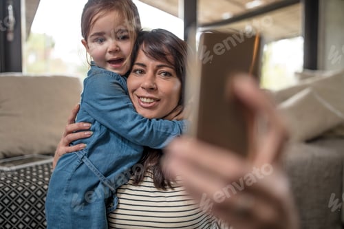 Preview: Mature woman taking smartphone selfie with daughter hugging her in living room