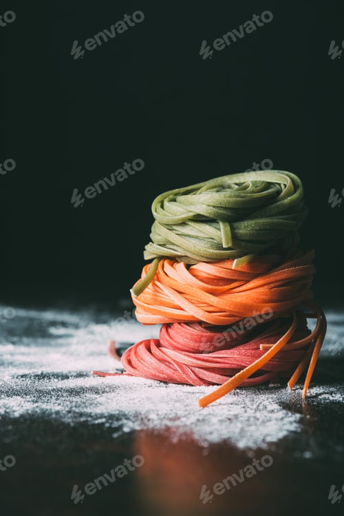 Preview: closeup shot of stack raw colorful tagliatelle pasta on table covered by flour