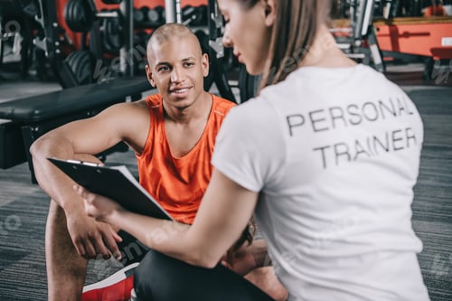 Preview: personal trainer showing clipboard to smiling african american sportsman sitting on floor