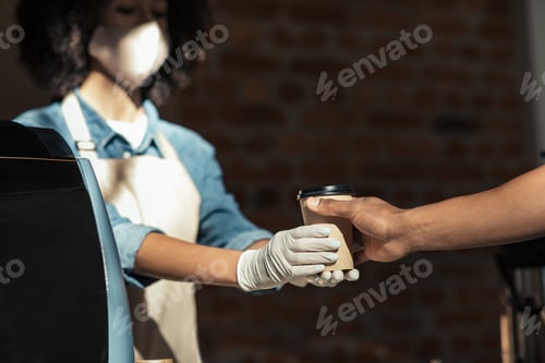 Preview: African american female cafe worker in face mask and gloves giving takeaway drink to client during